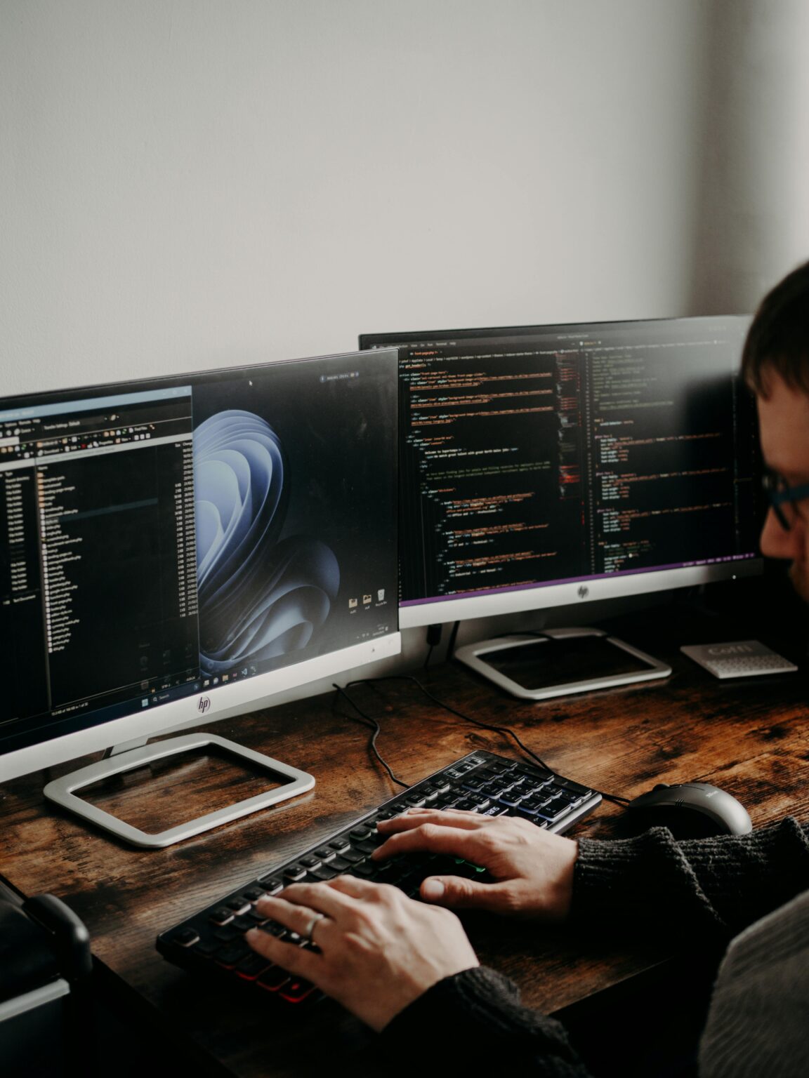 A man working on website design and coding at a home office with a dual monitor setup.